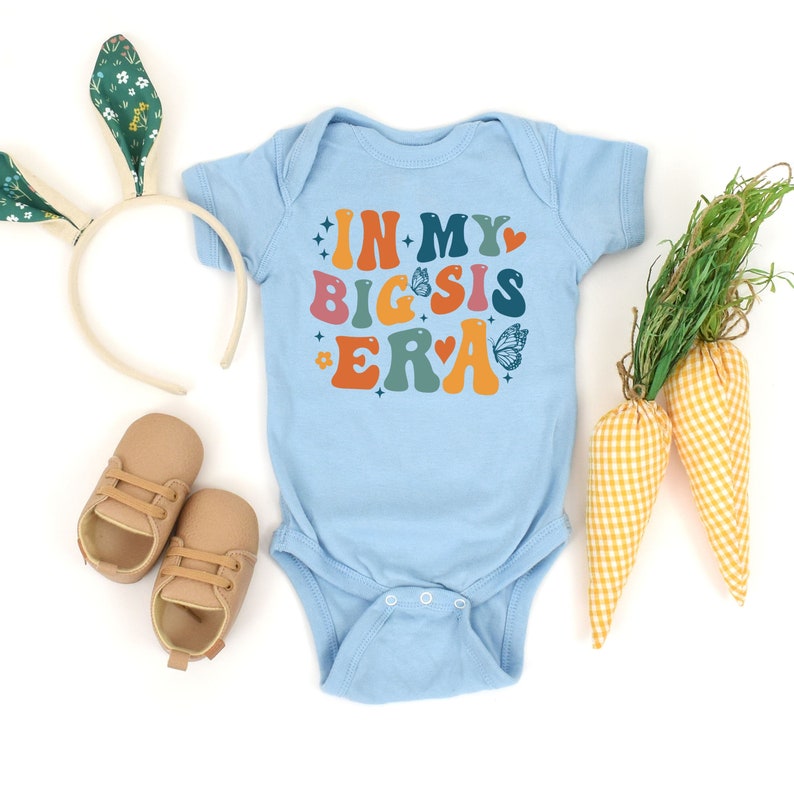 May include: A light blue baby onesie with the text "In My Big Sis Era" in colorful retro font. The onesie is decorated with hearts, butterflies, and stars.