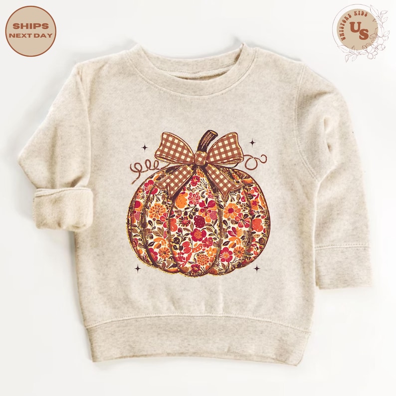 Puede incluir: Sudadera beige con dise&ntilde;o de calabaza floral. La calabaza est&aacute; llena de flores rojas, naranjas y amarillas, coronada con un lazo y tallo a cuadros marrones. El dise&ntilde;o incluye remolinos decorativos y peque&ntilde;as estrellas.