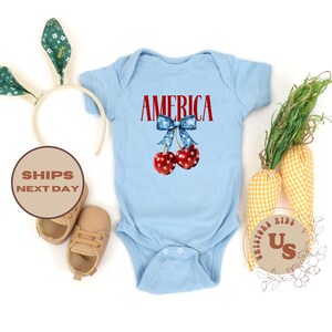 Op de afbeelding: Een lichtblauwe babybody met het woord "AMERICA" in rode letters boven een paar kersen met een blauwe strik. De kersen zijn rood met witte sterren. Een witte hoofdband met groene bloemenoren en beige babyschoenen staan ook op de foto.