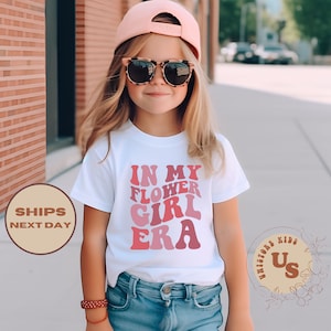 May include: A white t-shirt with the text "IN MY FLOWER GIRL ERA" in shades of pink. The shirt is worn by a young person wearing a pink cap and sunglasses.