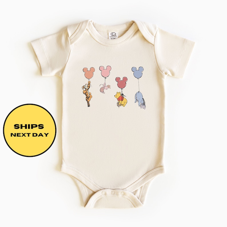 May include: A cream colored baby onesie with a graphic of Winnie the Pooh, Tigger, Piglet, and Eeyore holding onto Mickey Mouse shaped balloons. The balloons are in shades of pink, blue, yellow, and orange.
