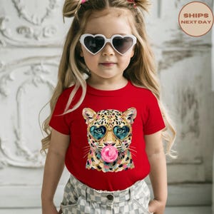 May include: A bright red short-sleeved t-shirt featuring a graphic print of a leopard wearing heart-shaped sunglasses and blowing a pink bubblegum bubble. The leopard illustration uses a colourful, textured paint-like style.