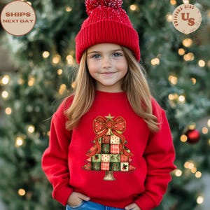 May include: A young person wearing a red sweatshirt and a matching red knit cap. The sweatshirt features a Christmas tree design with a large red bow and patchwork squares in green, red, and white. The background is a blurred Christmas tree.
