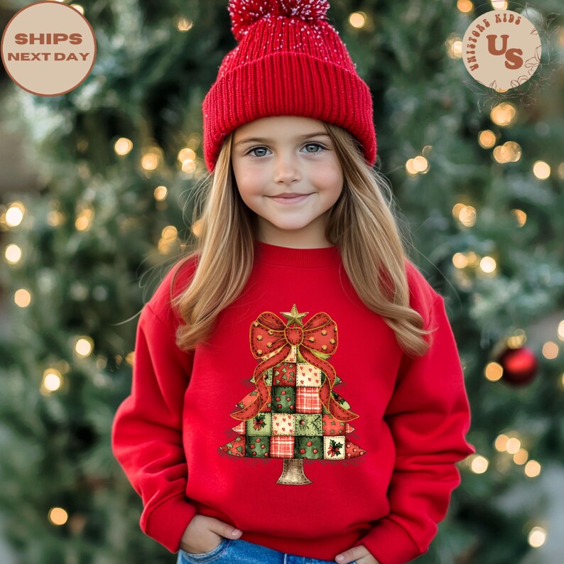 May include: A young person wearing a red sweatshirt and a matching red knit cap. The sweatshirt features a Christmas tree design with a large red bow and patchwork squares in green, red, and white. The background is a blurred Christmas tree.