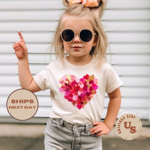 May include: A toddler wearing a white t-shirt with a pink and gold heart design made of small hearts. The shirt has the text "UniStore Kids" on the bottom right corner.