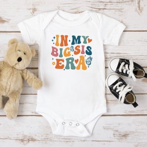 May include: White baby onesie with the text "IN MY BIG SIS ERA" in colorful retro font with butterflies and hearts.