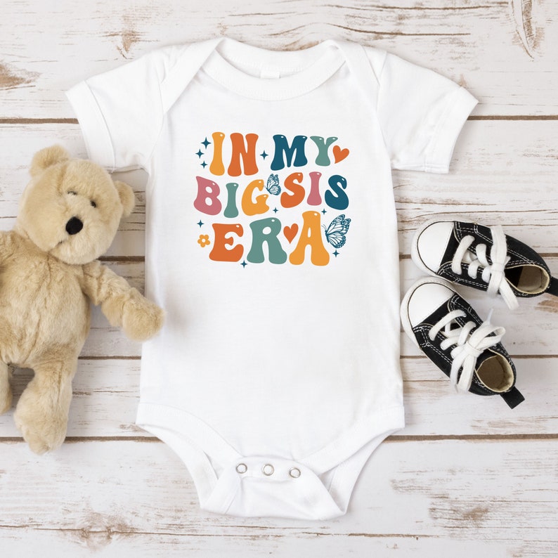 May include: White baby onesie with the text "IN MY BIG SIS ERA" in colorful retro font with butterflies and hearts.
