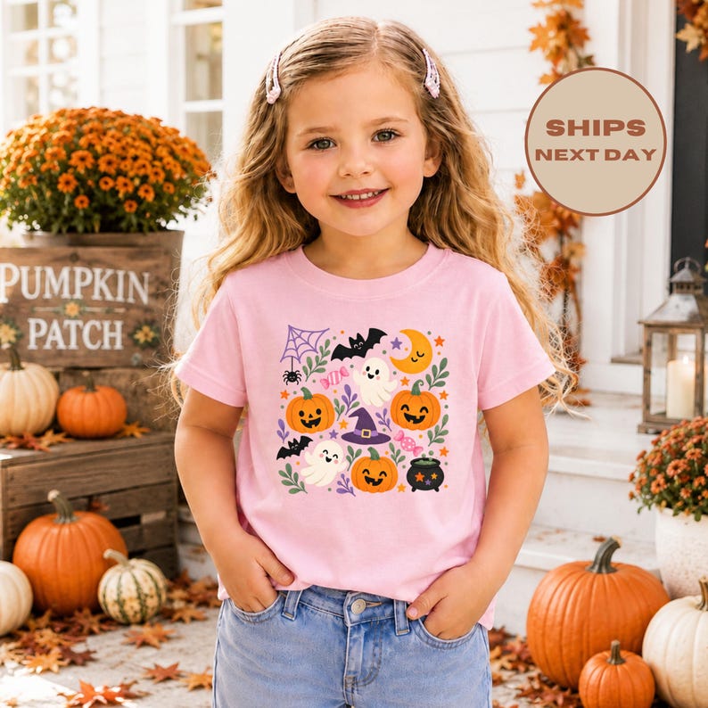 Puede incluir: Una camiseta de manga corta de color rosa claro con un estampado gr&aacute;fico colorido con tem&aacute;tica de Halloween. El dise&ntilde;o incluye calabazas sonrientes, fantasmas blancos, un sombrero de bruja morado, un murci&eacute;lago negro, una telara&ntilde;a y dulces.