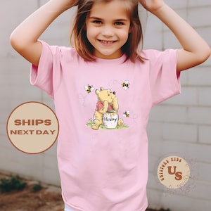 May include: Pink short-sleeved t-shirt featuring a cartoon Winnie the Pooh holding a pot of honey.  The shirt has a graphic of bees flying around Winnie the Pooh.