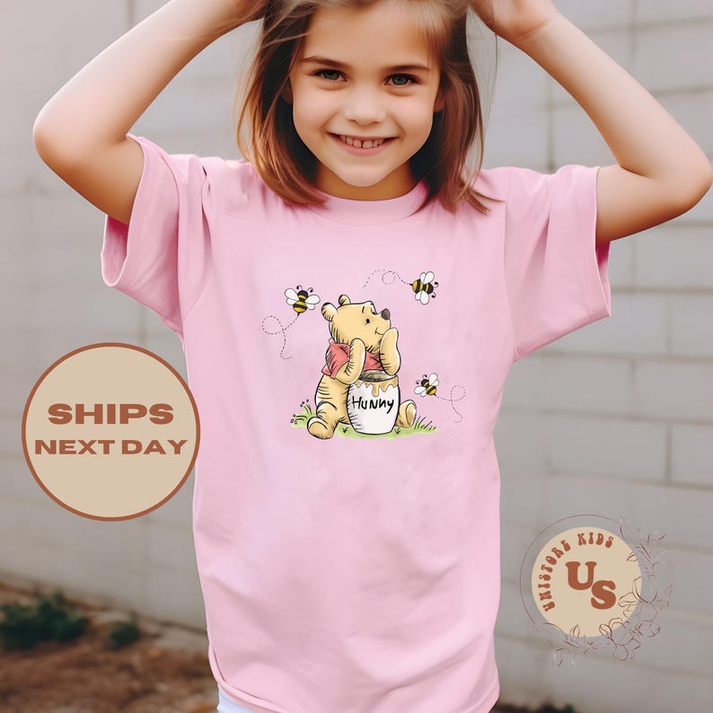 May include: Pink short-sleeved t-shirt featuring a cartoon Winnie the Pooh holding a pot of honey.  The shirt has a graphic of bees flying around Winnie the Pooh.