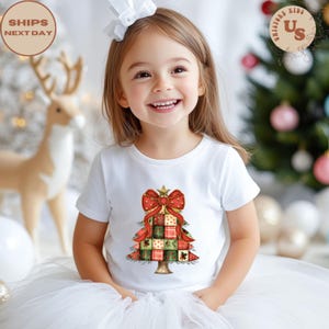 May include: A young child wearing a white t-shirt with a Christmas tree design. The tree is made of red, green, and gold squares, topped with a red bow and star. The shirt is part of a holiday collection.