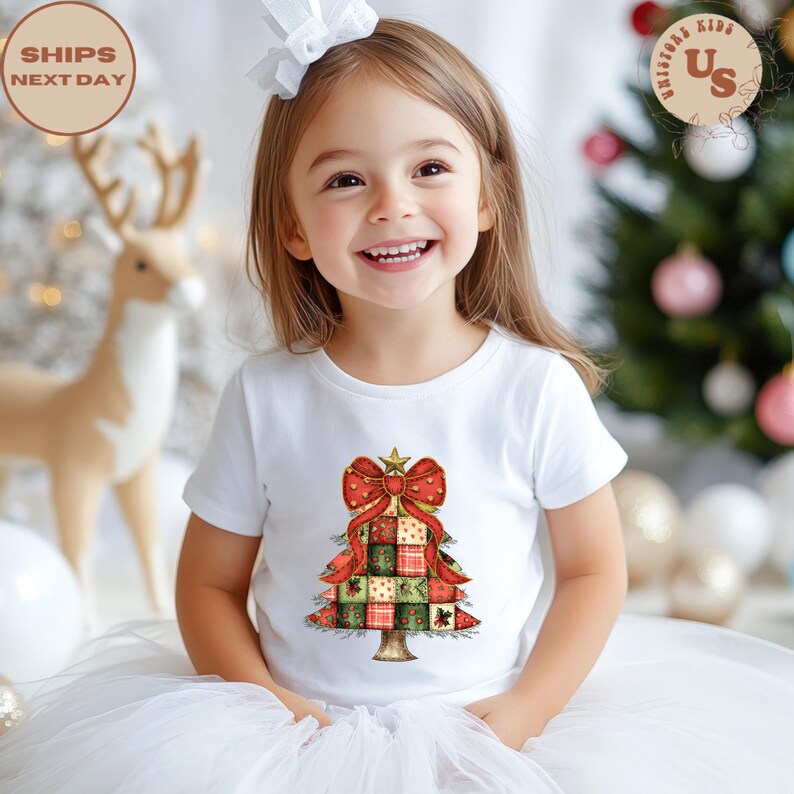 May include: A young child wearing a white t-shirt with a Christmas tree design. The tree is made of red, green, and gold squares, topped with a red bow and star. The shirt is part of a holiday collection.