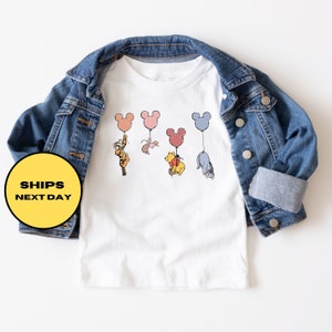 May include: White t-shirt with a graphic of Winnie the Pooh, Piglet, Tigger, and Eeyore holding onto Mickey Mouse balloons. The balloons are different colors, including pink, blue, and yellow.