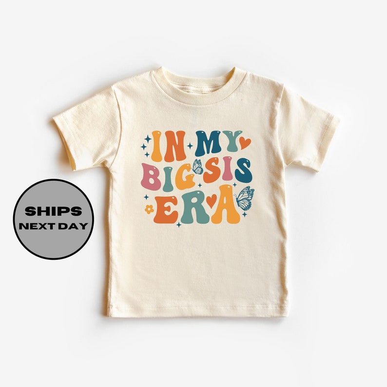 May include: A light beige short-sleeved t-shirt with the text "IN MY BIG SIS ERA" in colorful retro-style lettering with butterflies and stars.