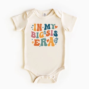 May include: A cream-colored baby onesie with short sleeves features a colorful graphic that reads "In My Big Sis Era" with butterflies and stars.