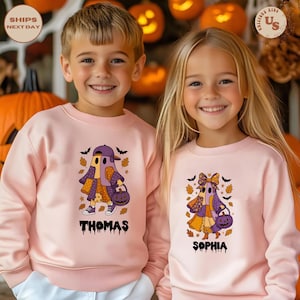May include: Two children in matching pink Halloween sweatshirts. Each sweatshirt features a ghost character with a hat and a pumpkin bag. The boy's shirt says "THOMAS", the girl's says "SOPHIA". Autumn leaves and bats surround the ghosts.