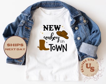 May include: White t-shirt with a brown cowboy boot and hat graphic. The text "NEW cowboy in TOWN" is printed on the shirt.