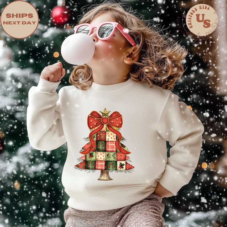 May include: A cream-colored sweatshirt featuring a Christmas tree design. The tree is composed of red, green, and gold patchwork squares, topped with a red bow and a gold star. The image includes the text "SHIPS NEXT DAY".