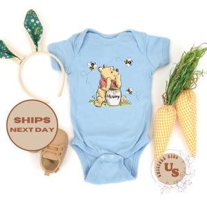 May include: A light blue baby onesie with a Winnie the Pooh illustration. Pooh is holding a jar of honey and surrounded by bees. The onesie has a white snap closure.