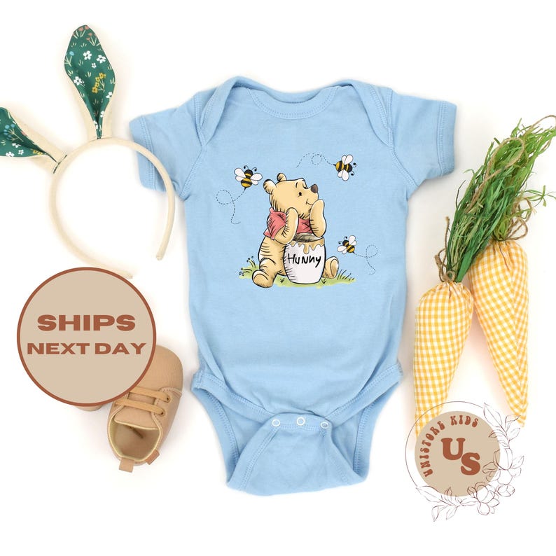 May include: A light blue baby onesie with a Winnie the Pooh illustration. Pooh is holding a jar of honey and surrounded by bees. The onesie has a white snap closure.