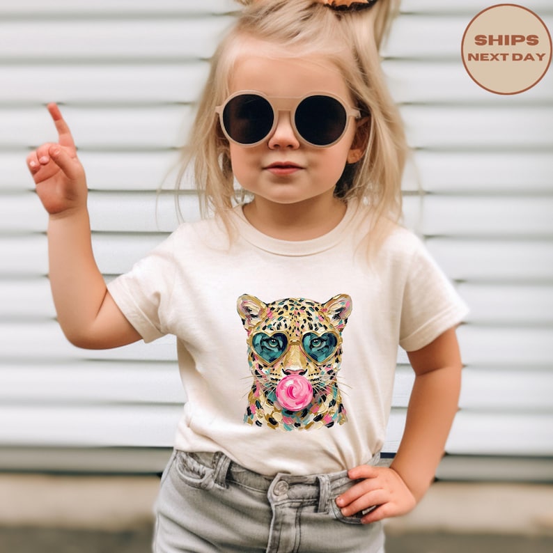 May include: A cream-coloured short-sleeve t-shirt featuring a graphic print of a leopard wearing heart-shaped sunglasses and blowing a pink bubblegum bubble. The leopard illustration uses a colourful, textured paint-stroke style.