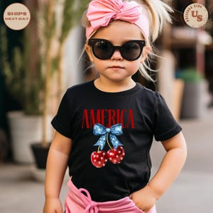 Op de afbeelding: Een zwart t-shirt met het woord "AMERICA" in het rood en een afbeelding van kersen met een blauwe strik. De kersen zijn rood met witte sterpatronen. Het kind draagt een zonnebril en een roze haarband.