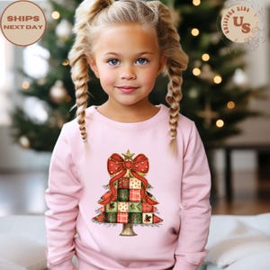 May include: A light pink sweatshirt featuring a Christmas tree design. The tree is composed of patchwork squares in red, green, and beige, topped with a red bow and a gold star. The sweatshirt is for children.