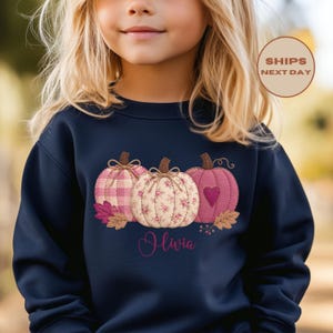 May include: A navy blue long-sleeve sweatshirt featuring a graphic of three pumpkins in shades of pink. The left pumpkin has a pink gingham pattern, the center has a floral print, and the right has a heart design. The name Olivia is printed below.