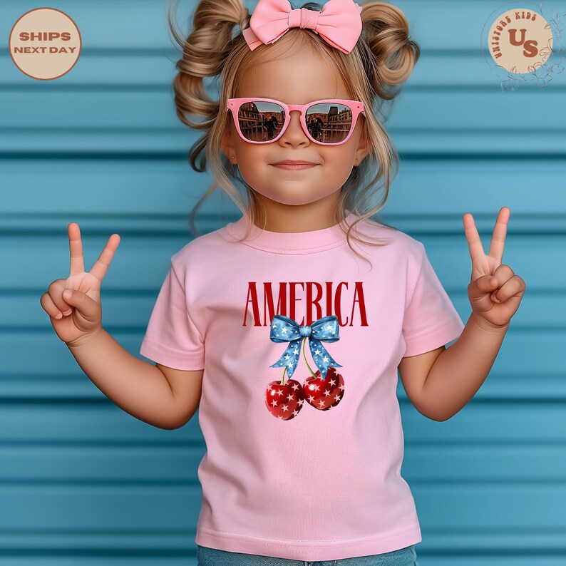 Op de afbeelding: Een roze t-shirt met het woord "AMERICA" in rode letters en een afbeelding van kersen met een blauwe strik. De kersen hebben een rood-wit sterrenpatroon. Het kind draagt een roze zonnebril en een roze strik.