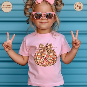 Puede incluir: Una camiseta rosa con un dise&ntilde;o de calabaza floral y un lazo a cuadros marr&oacute;n. La camiseta la lleva un ni&ntilde;o que lleva gafas de sol rosas y un lazo rosa. El fondo es una pared azul.
