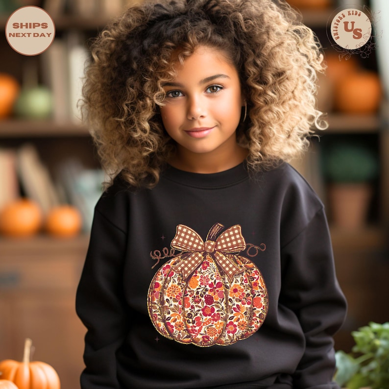 Puede incluir: Una sudadera negra con un dise&ntilde;o de calabaza floral y un lazo de cuadros. La calabaza est&aacute; llena de flores naranjas, rojas y amarillas. La sudadera la lleva una persona joven con el pelo rizado.