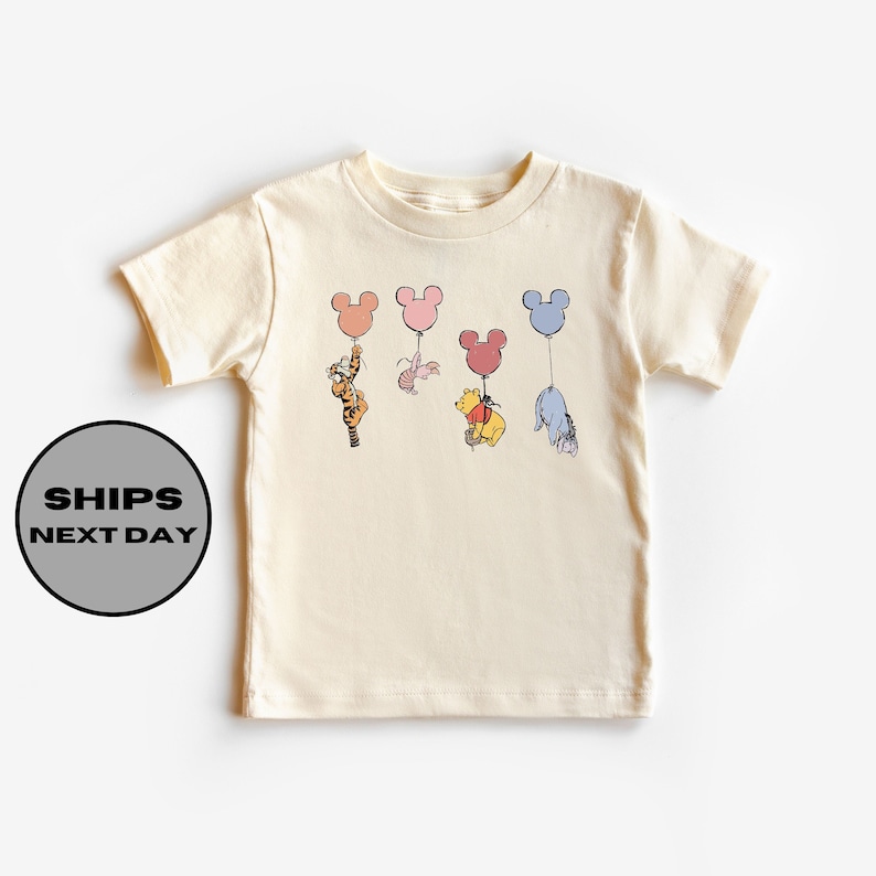May include: A cream colored t-shirt with a graphic of Winnie the Pooh, Tigger, Piglet, and Eeyore holding onto Mickey Mouse shaped balloons. The balloons are in shades of pink, blue, and yellow.