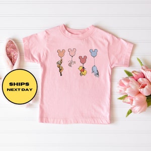 May include: Pink toddler t-shirt with a graphic of Winnie the Pooh, Tigger, Piglet, and Eeyore holding onto Mickey Mouse balloons.