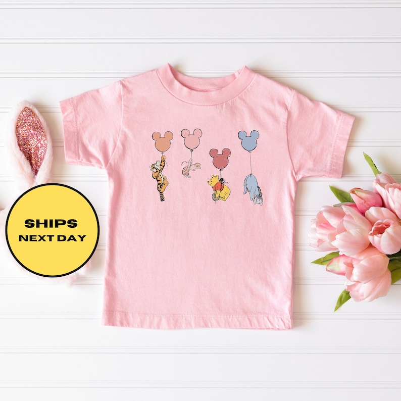 May include: Pink toddler t-shirt with a graphic of Winnie the Pooh, Tigger, Piglet, and Eeyore holding onto Mickey Mouse balloons.