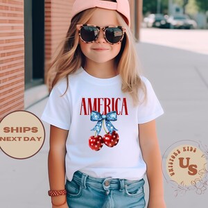 Op de afbeelding: Een wit t-shirt met het woord "AMERICA" in rode letters en een blauwe strik met rood-witte kersen. Het t-shirt wordt gedragen door een jong persoon met een zonnebril en een roze pet.