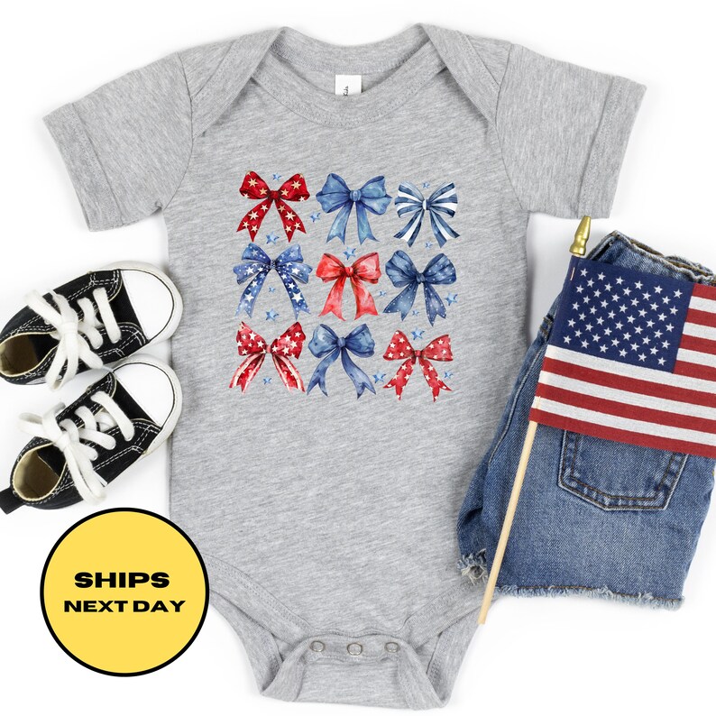 May include: A gray baby onesie with a pattern of red, white, and blue bows. The bows are arranged in a grid pattern and have a watercolor effect. The onesie is on a white background.