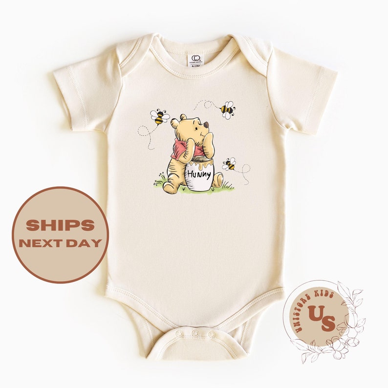 May include: A cream colored baby onesie with a cartoon image of Winnie the Pooh holding a jar of honey.  The text on the jar reads "Hunny".  There are three bees flying around Winnie the Pooh.