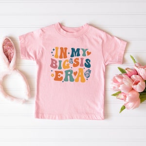 May include: Pink t-shirt with the text "In My Big Sis Era" in colorful retro letters with butterflies and stars.