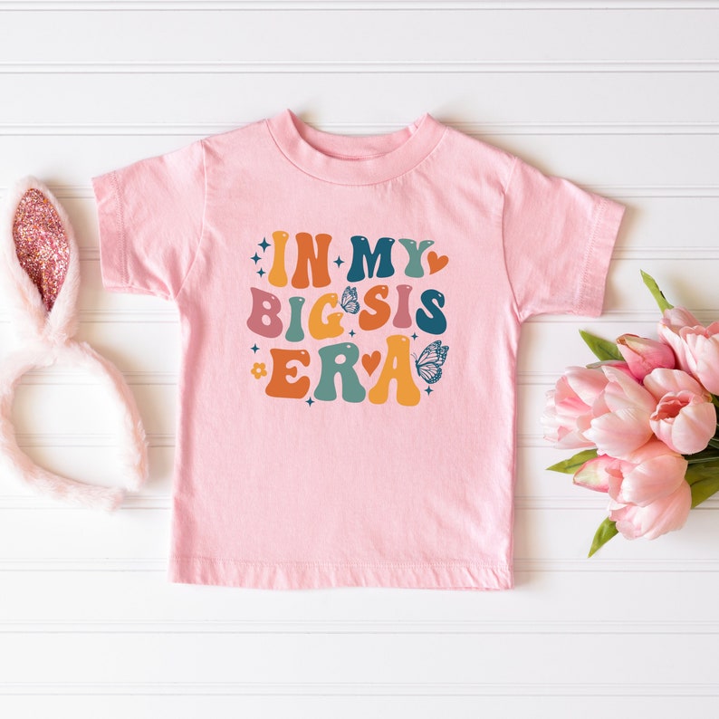May include: Pink t-shirt with the text "In My Big Sis Era" in colorful retro letters with butterflies and stars.