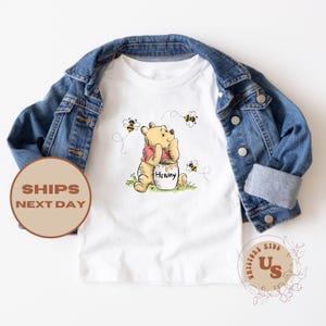 May include: White t-shirt with a cartoon image of Winnie the Pooh holding a honey pot. The shirt has a red, white, and yellow color scheme. There are bees flying around Winnie the Pooh.