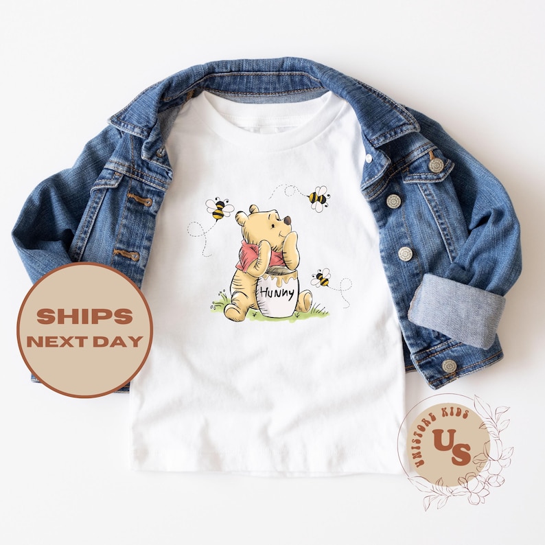 May include: White t-shirt with a cartoon image of Winnie the Pooh holding a honey pot. The shirt has a red, white, and yellow color scheme. There are bees flying around Winnie the Pooh.