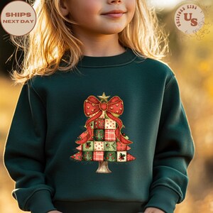May include: A dark green sweatshirt featuring a Christmas tree design. The tree is composed of patchwork squares in red, green, and white, adorned with a red bow and a gold star. The text "SHIPS NEXT DAY" is visible.