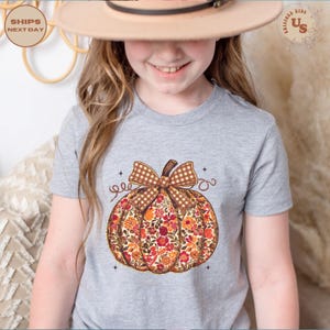 Puede incluir: Una camiseta gris con un dise&ntilde;o de calabaza floral y un lazo a cuadros marr&oacute;n. La calabaza est&aacute; llena de flores naranjas, rojas y amarillas. La camiseta es de talla infantil.