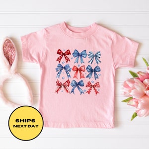 May include: Pink toddler t-shirt with a design of red, white, and blue bows with stars.