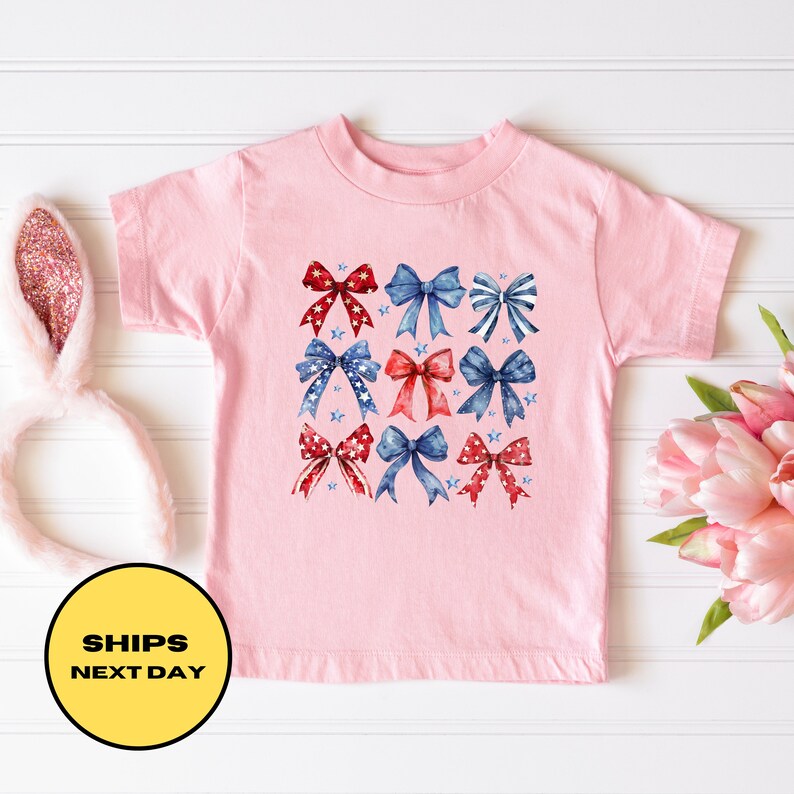 May include: Pink toddler t-shirt with a design of red, white, and blue bows with stars.