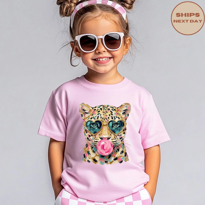 May include: A light pink t-shirt featuring a graphic print of a leopard wearing heart-shaped sunglasses and blowing a pink bubblegum bubble. The leopard has a spotted coat with gold, black, and teal accents, and the shirt has a crew neckline.