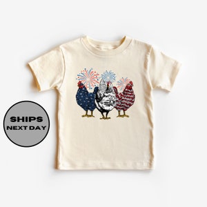 May include: Cream-colored t-shirt featuring a graphic of three chickens with patriotic designs and fireworks. The chickens are colored with red, white, and blue patterns. The text "SHIPS NEXT DAY" is in a black circle on the left side of the shirt.