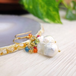 Peut inclure: Boucles d'oreilles pendantes dorées avec de grosses perles blanches irrégulières et des grappes de pierres précieuses colorées. Les boucles d'oreilles ont une fermeture à crochet. Elles sont présentées sur une surface claire avec un fond gris et jaune.