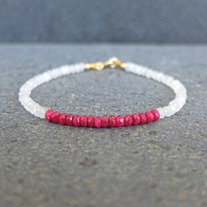 Può includere: Un braccialetto delicato realizzato con perline bianche e rosse. Il braccialetto presenta una fila di piccole perline rosso vivo al centro, circondate da perline bianche.