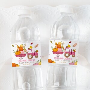 Puede incluir: Dos botellas de agua de plástico transparente con etiquetas que muestran un tractor rosa transportando calabazas y hojas de otoño. Las etiquetas también incluyen el texto "Emily and James's Baby Shower".
